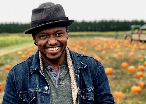 Omi is standing in front of a pumpkin patch, smiling. They have dark hat, a jean jacket and cozy sweater.
