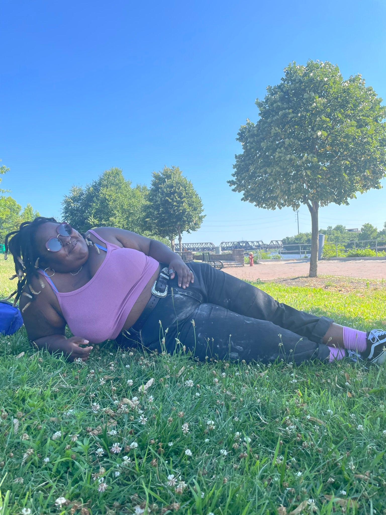 Ronnie is laying in some grass in the shade, they are wearing black and white vans, pink socks, black denim jeans, pink spaghetti strap tank top and some of their stomach is showing, they are smiling and wearing heart shaped sunglasses