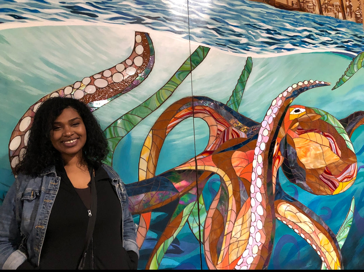 Tasha is smiling and standing in front of a colorful mural of an octopus. She is wearing a jean jacket with a black sweater underneath. She has dark brown hair which is loose and curly and she has brown skin.