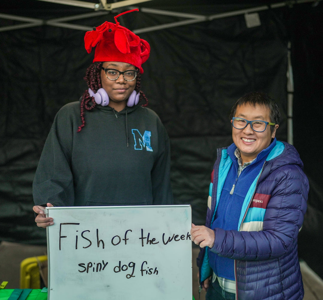 Talia and a student wearing a lobster hat hold a handwritten sign saying "Fish of the Week Spiny Dogfish" at a fishstand. This is a still from the Hope in the Water segment