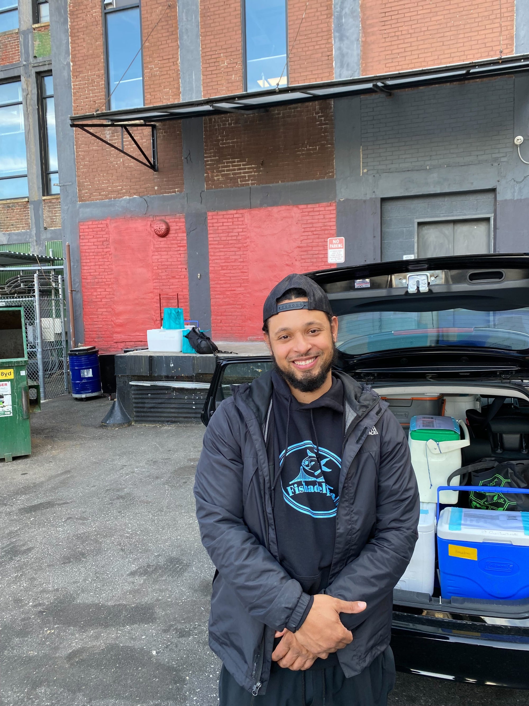 Tino stands in front of an open car truck with coolers full of fish. He is smiling and wearing a Fishadelphia sweatshirt