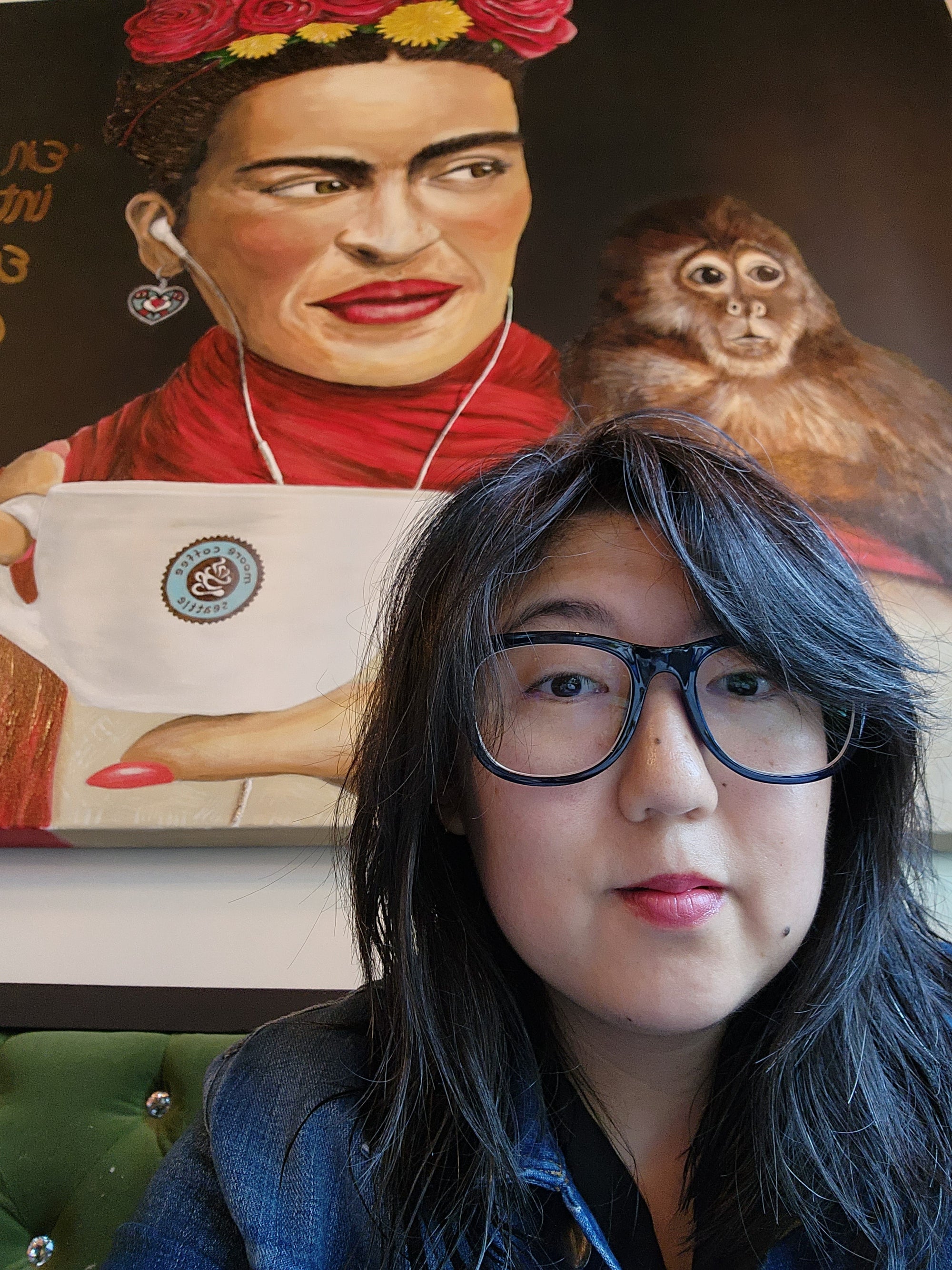 Victoria sits in front of a painting of Frida Kahlo. Victoria has long dark hair, dark glasses, and red lips.