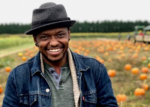 Omi is standing in front of a pumpkin patch, smiling. They have dark hat, a jean jacket and cozy sweater.