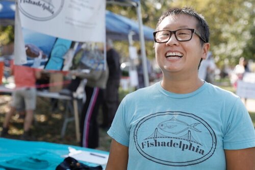 Talia is outside at a Fishadelphia fishstand, wearing a blue Fishadelphia logo t-shirt and laughing. She has short dark hair and glasses.